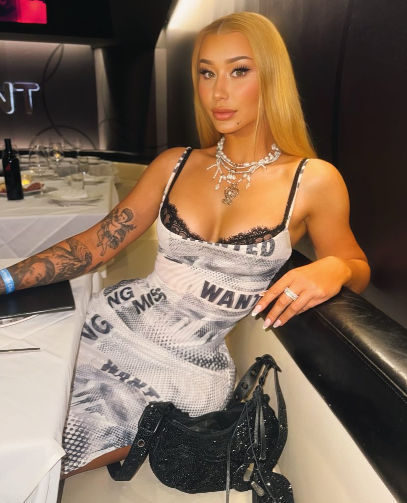 Rapper Iggy Azalea sitting at a restaurant table wearing a "WANTED" print dress and layered pearl necklaces, presenting a glamorous aesthetic popular with fans of her music and her Iggy Azalea OnlyFans account.
