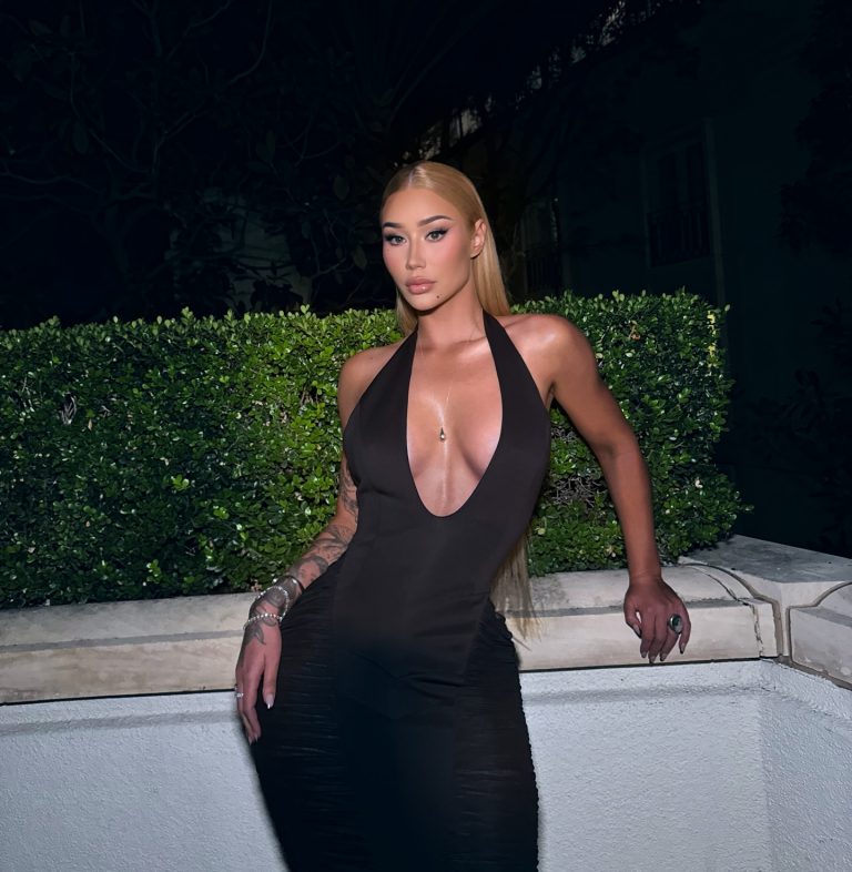 Iggy Azalea standing on a balcony overlooking a garden, wearing a sleek black strapless dress with a sheer midsection panel and holding a metallic clutch, reflecting the high-fashion imagery connected to her Iggy Azalea OnlyFans brand.