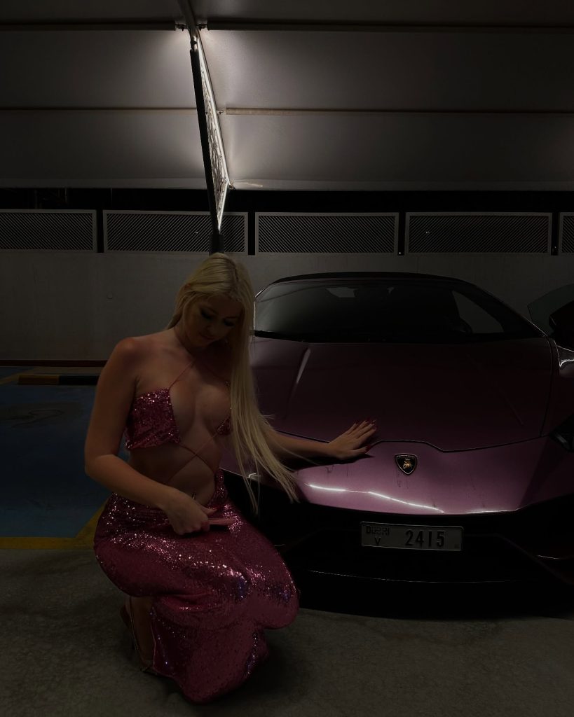 Popular Greek Onlyfans star Iro Kasouni kneeling in a dimly lit garage next to a luxury purple Lamborghini, wearing a sparkling pink sequin two-piece dress.