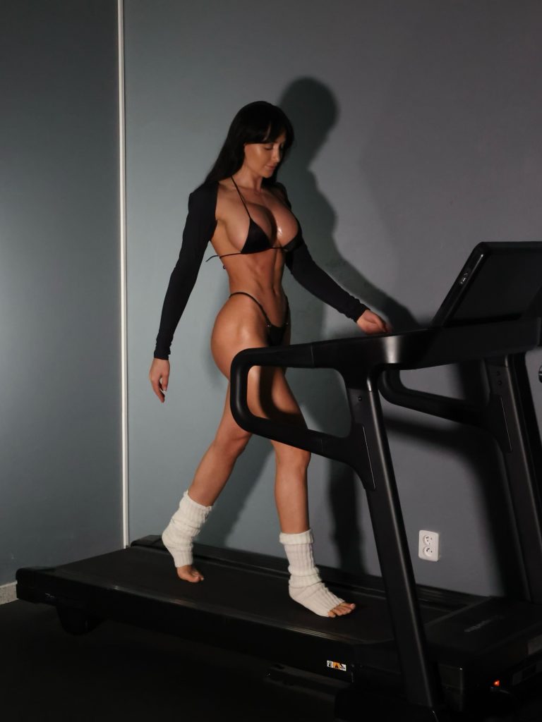 Kristýna Sixtová walking on a treadmill in a fitness studio, wearing a black string bikini and white leg warmers.