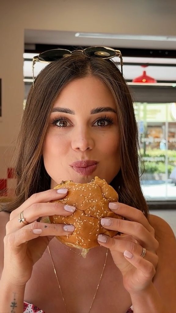 A close-up profile shot of Czech OnlyFans star Little Caprice holding and eating a hamburger in a restaurant setting.