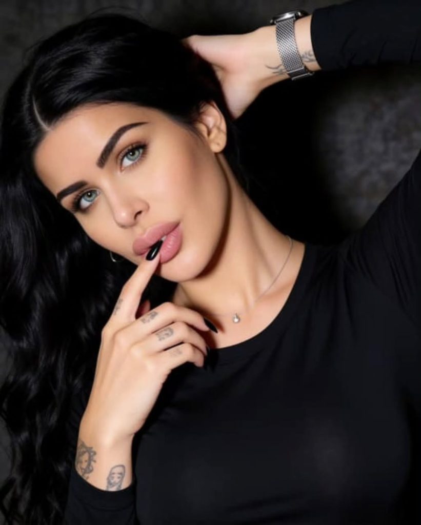 A close-up portrait of a Maria Saatsaki with dark hair and striking green eyes, holding a finger to her lips while posing in a black top against a dark background.