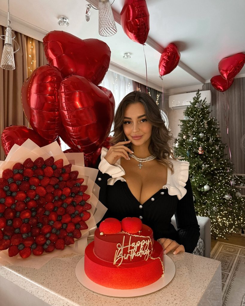 Glamour influencer Marina Nabokina celebrating her birthday indoors with a two-tier red cake, a large strawberry bouquet, and red heart-shaped balloons, with a Christmas tree in the background.