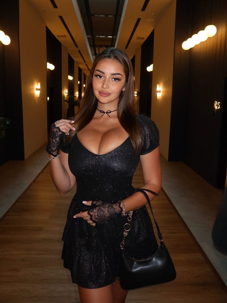 Marina Nabokina, a Russian OnlyFans model, posing in a dimly lit hallway wearing a sparkling black dress, lace fingerless gloves, and a black choker with a bow.