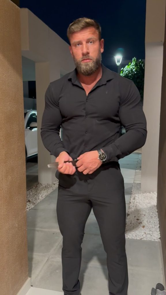 Irish OnlyFans creator Matty Gilbert in a fitted black button-down shirt and black trousers, adjusting his belt in an outdoor corridor.