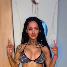 Irish OnlyFans creator Meghan O'Neill posing with dark dreadlocks, a spiderweb pattern bikini top, and intricate neck and arm tattoos while pointing upward.