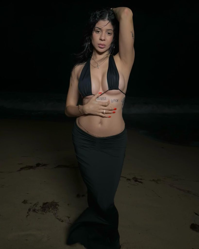 Nicole Castillo Puerto Rican OnlyFans posing on a beach at night with dark ocean waves behind her. She wears a black halter bikini top paired with a long, low-rise black maxi skirt and a thin gold waist chain.