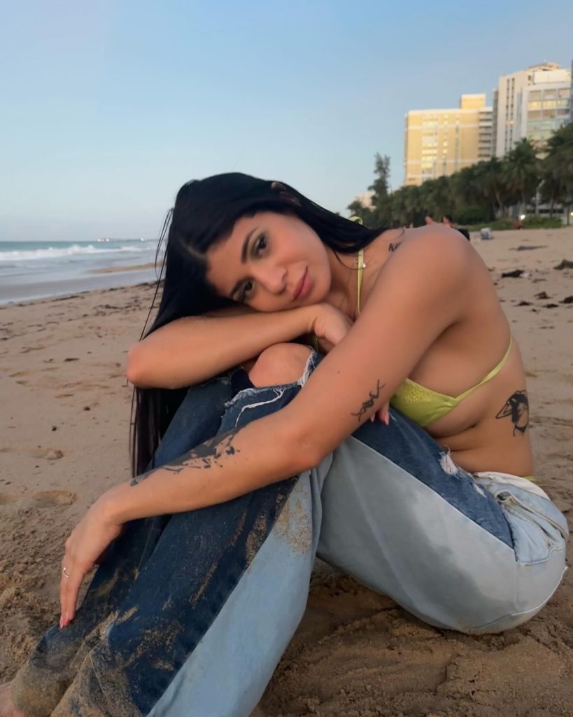 Puerto Rican OnlyFans creator Nicole Castillo sitting on a sandy beach at sunset, resting her head on her arms. She is wearing a light green bikini top and distressed, multi-toned blue jeans.