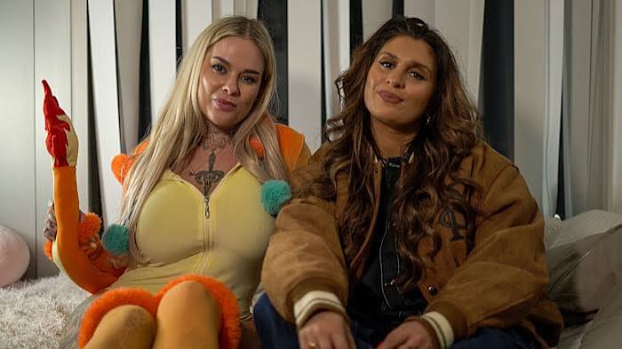 Two creators from an OnlyFans Agency sitting together on a white rug; one woman wears a yellow and orange outfit with pom-poms while the other wears a brown varsity-style jacket.