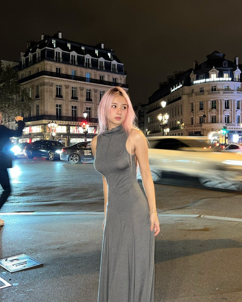 A candid, medium close-up shot of a woman with pink hair looking to her left while walking on a busy Parisian street at night. She is wearing a long, grey sleeveless knit dress
