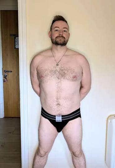 Full-body portrait of bearded Irish OnlyFans model Prince Trek standing against a plain wall in black athletic briefs.