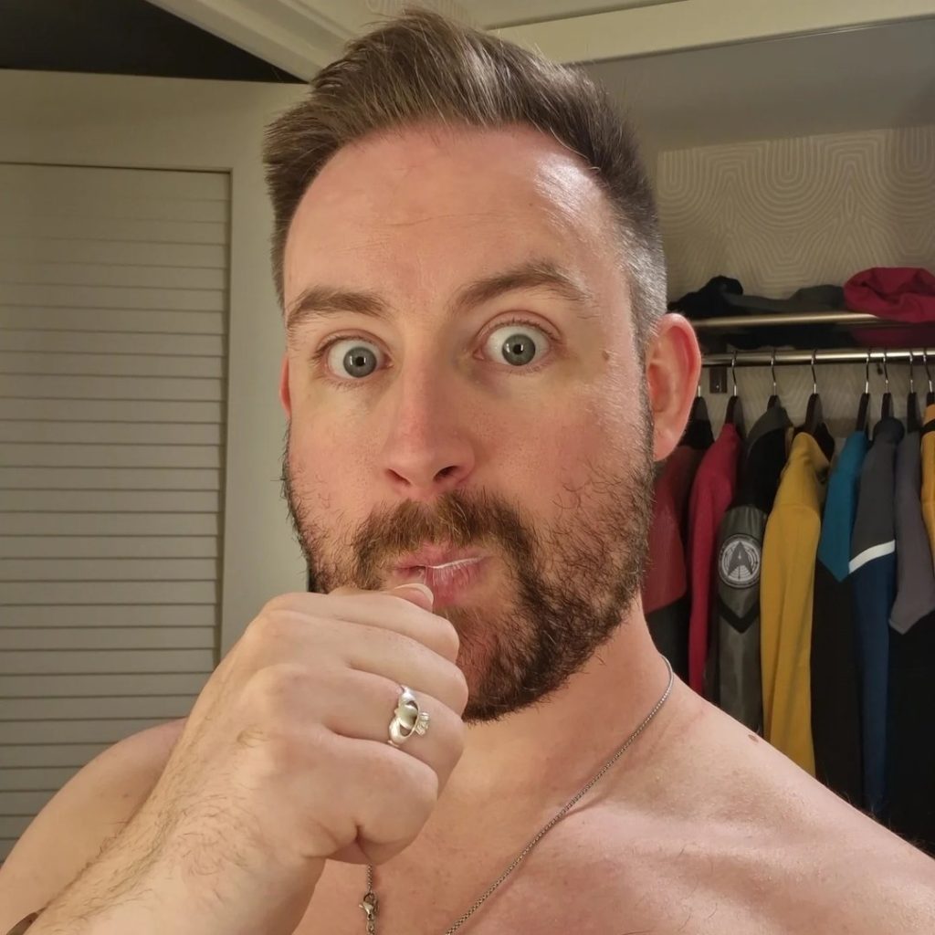 Close-up selfie of bearded Irish OnlyFans model Sean Ferrick with an expressive wide-eyed look, holding a ring to his mouth with a clothing rack of colorful jackets in the background.