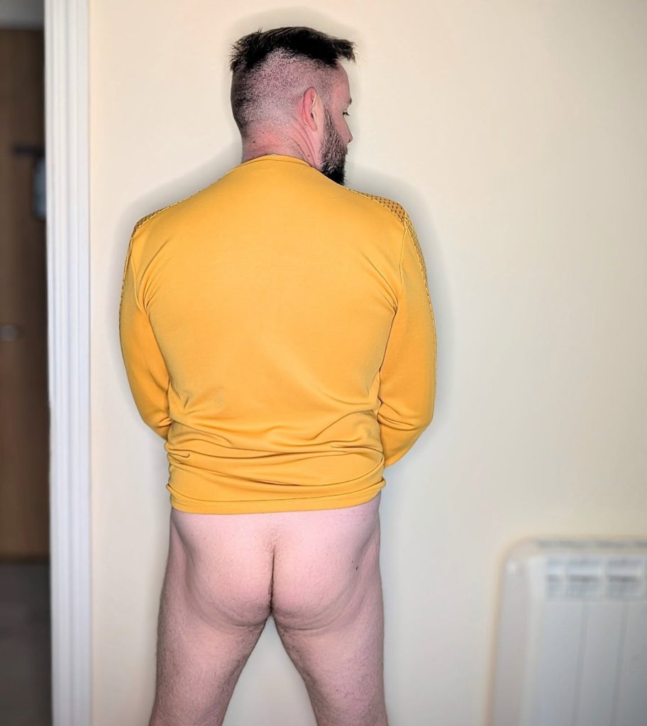 Irish content creator Sean Ferrick pictured from behind, wearing a long-sleeved gold shirt while exposed from the waist down against a plain wall.