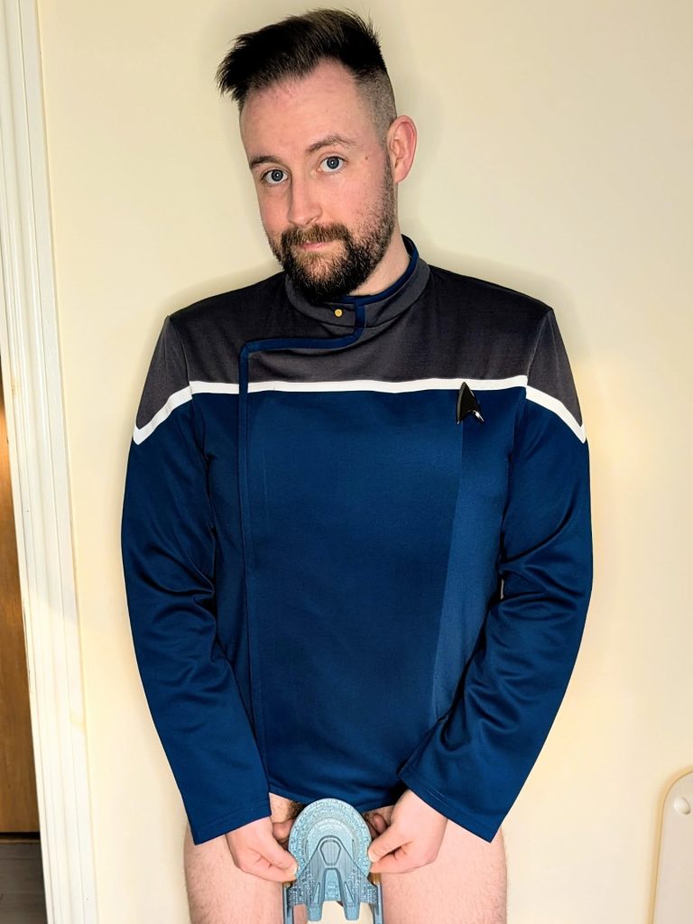 Irish OnlyFans influencer Sean Ferrick posing in a blue and gray Star Trek uniform top, holding a toy starship model to cover himself from the waist down.