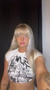 A vertically oriented, slightly blurry selfie of a young woman with platinum blonde hair and straight-cut bangs. She is looking forward with a neutral expression. She is wearing a white crop top t-shirt featuring a black graphic print text design. Part of a tattoo is visible on her lower left forearm. The background consists of a dark, solid curtain or wall on the left and a plain white wall on the right.