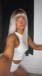 A vertical, slightly low-angle selfie of a young woman with long platinum blonde hair and straight bangs, captured mid-pose. She is leaning forward toward the camera, looking straight ahead. She is wearing a sleeveless white high-neck crop top and matching white shorts. The background features a dark curtain and a white baseboard along the wall.