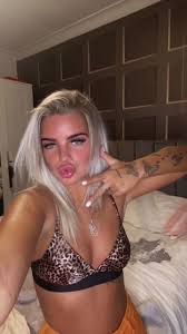 A vertical selfie of a young woman with platinum blonde hair looking into the camera while making a pouty face and holding up a two-finger peace sign next to her face. She has a visible tattoo of butterflies on her left forearm. She is wearing a leopard-print bralette and orange bottoms. The background shows a bedroom setting with a padded brown headboard and a white wardrobe.
