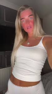 A vertical, slightly soft-focus indoor selfie of a young woman with long platinum blonde hair parted down the middle. She has a noticeably warm or sun-kissed complexion and is looking at the camera with her mouth slightly open. She is wearing a simple white ribbed camisole tank top and white bottoms. A small silver cross necklace hangs around her neck. The background shows a dimly lit domestic room.