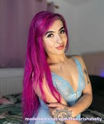 A casual indoor portrait of Irish OnlyFans model Shelly smiling gently at the camera. Her bright purple-pink hair stands out against a soft bedroom background as she poses in light blue lace lingerie, revealing a detailed sun tattoo on her forearm.