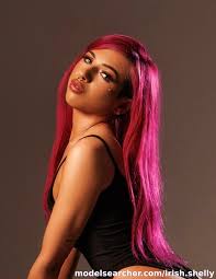 A close-up studio portrait of Irish OnlyFans creator Shelly looking thoughtfully over her shoulder. She features vibrant, long magenta hair, a nose ring, and subtle facial gems, posing in a simple black tank top against a warm, neutral studio backdrop.
