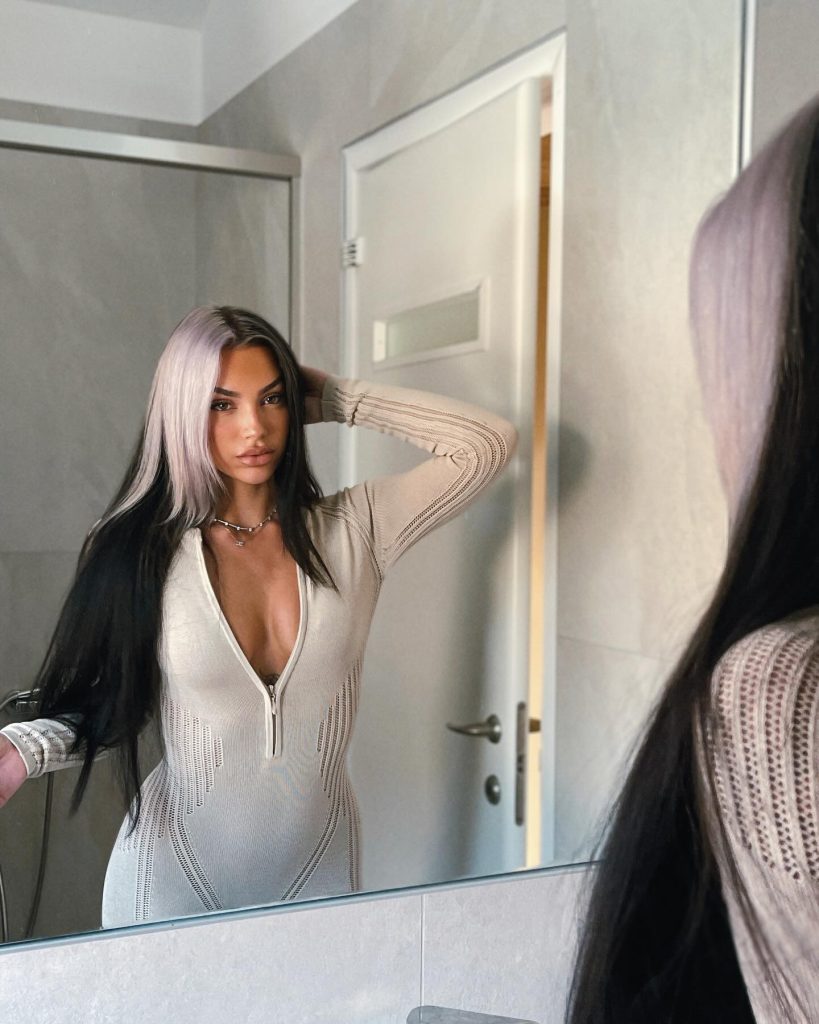 A mirror selfie of Greek Onlyfans personality Sophia Rizou in a bright bathroom, wearing a long-sleeved beige jumpsuit unzipped at the front while running a hand through her split-dyed blonde and black hair.
