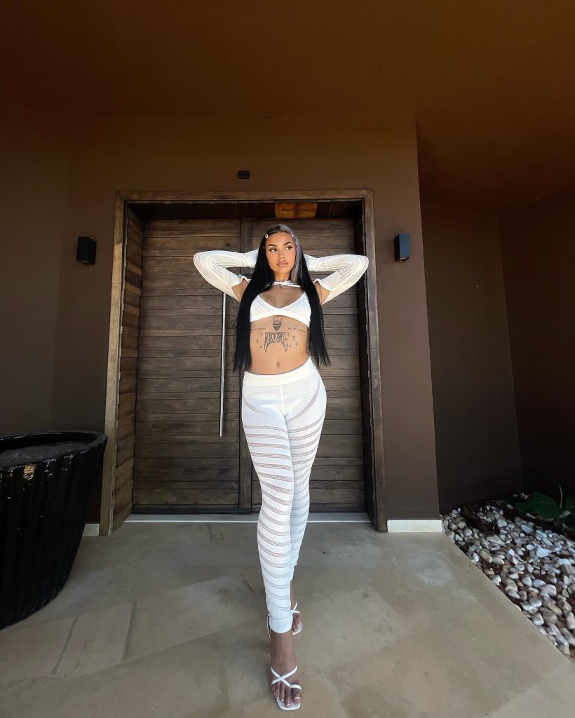 Sophia Rizou posing in front of large wooden doors, wearing a white two-piece crop top and leggings outfit with patterned cutouts, showcasing her midriff tattoos and half-blonde, half-black hair.