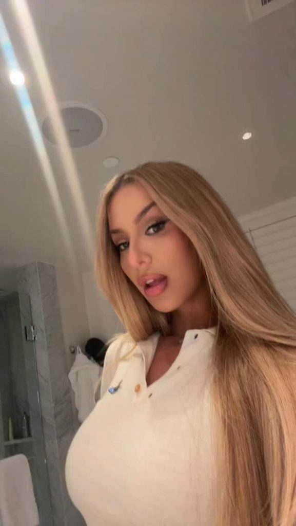 A low-angle portrait of Tana Mongeau indoors with long blonde hair, looking toward the camera in a cream-colored collared shirt