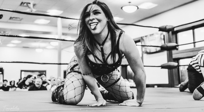 Black and white action shot of Vanilla Vargas crawling in a wrestling ring, smiling and sticking her tongue out toward the audience. She is wearing a strapped leather top and fishnet tights.