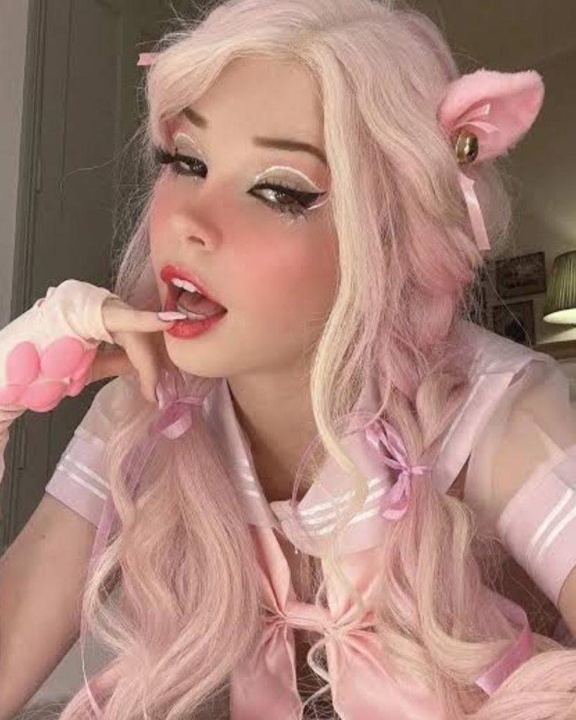 Belle Delphine OnlyFans influencer posing in a pastel pink anime-inspired cosplay outfit with matching pink hair, plush cat ears with bells, and dramatic white graphic eyeliner.