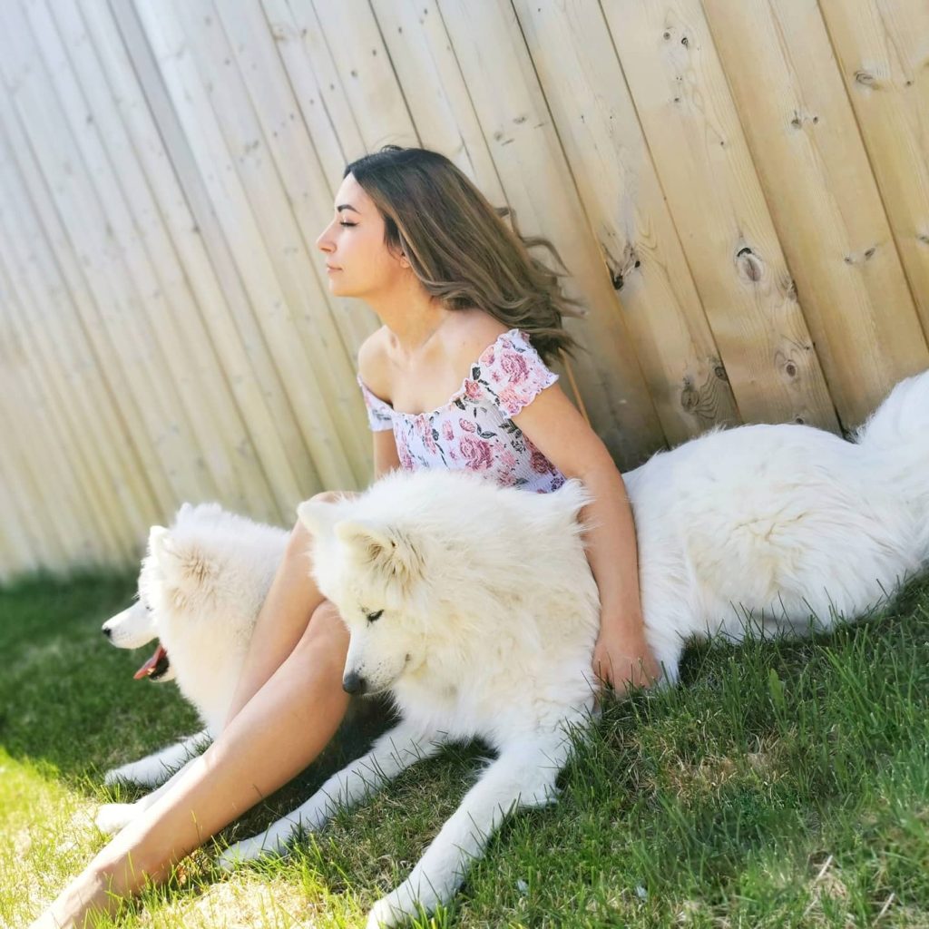 Alinity sits peacefully on a grassy lawn against a light wooden fence, wearing a floral off-the-shoulder top while petting two large, fluffy white Samoyed dogs, highlighting an intimate lifestyle look