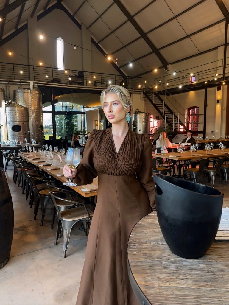 Elegant portrait of Amber Brits in a modern winery, holding a glass of white wine, with large metal fermentation tanks in the background.