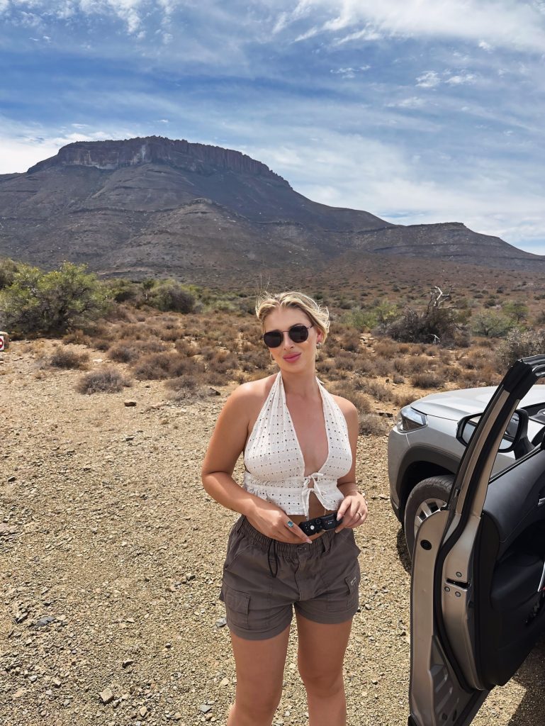 Candid travel shot of Amber Brits wearing sunglasses and a white eyelet halter top in a dry, mountainous South African landscape next to a silver SUV.