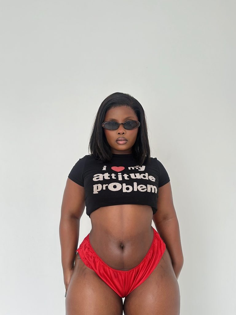 Full-body studio shot of Anelay wearing oval sunglasses, a black "attitude problem" crop top, and red bottoms against a white wall.