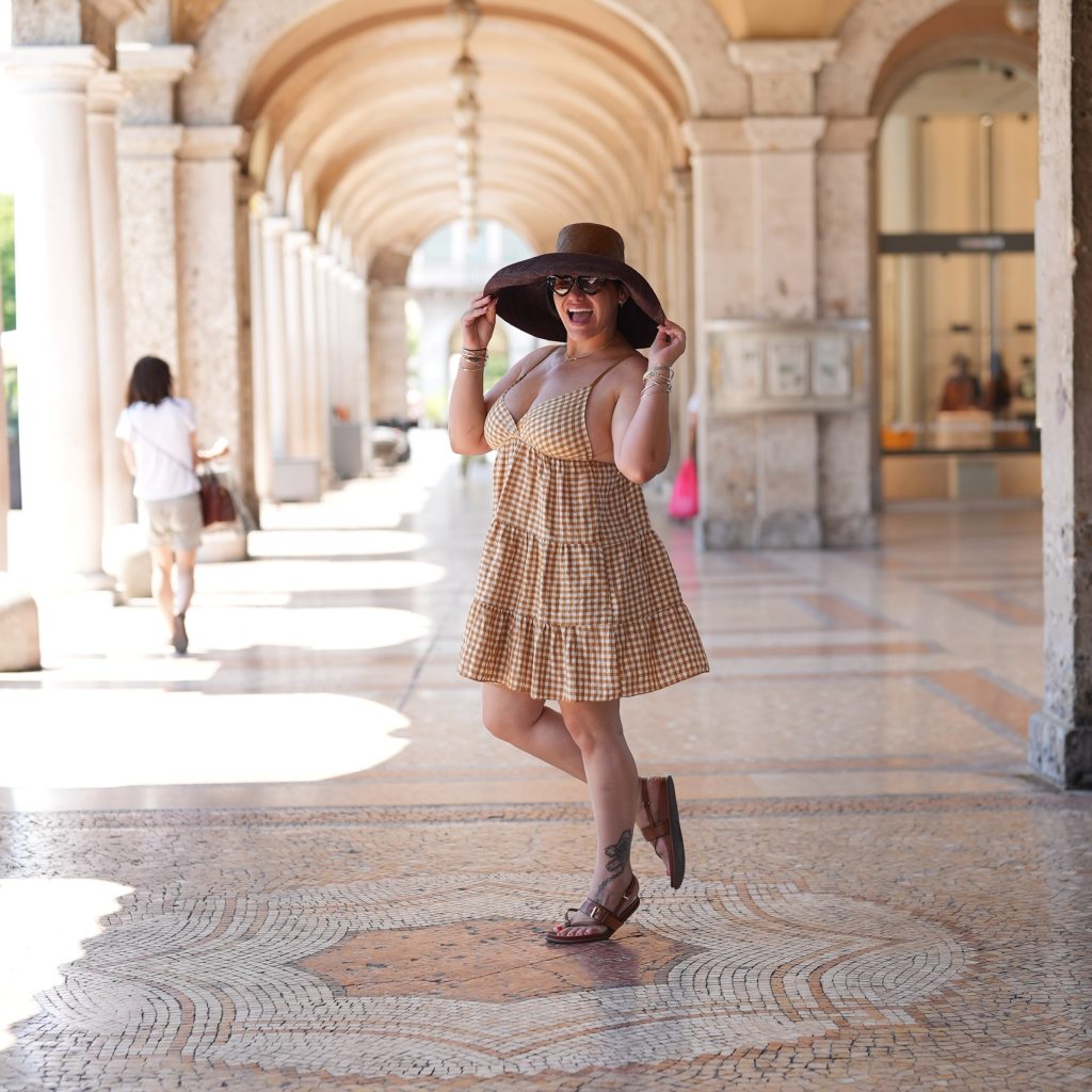 A cheerful, full-length shot of a curvy woman posing playfully in an ornate Italian-style colonnade, holding the wide brim of her straw hat while wearing a checkered sundress. This image may represent content from a Cuban OnlyFans creator.