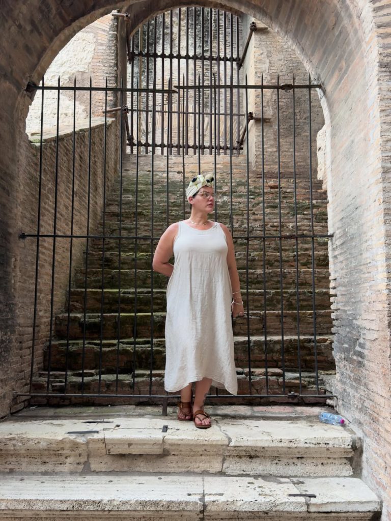 A full-length photograph of a woman wearing a white linen dress, headwrap, and sandals, standing before a rusted iron gate and stone staircase. Context implies this image may be associated with content from a Cuban OnlyFans profile.