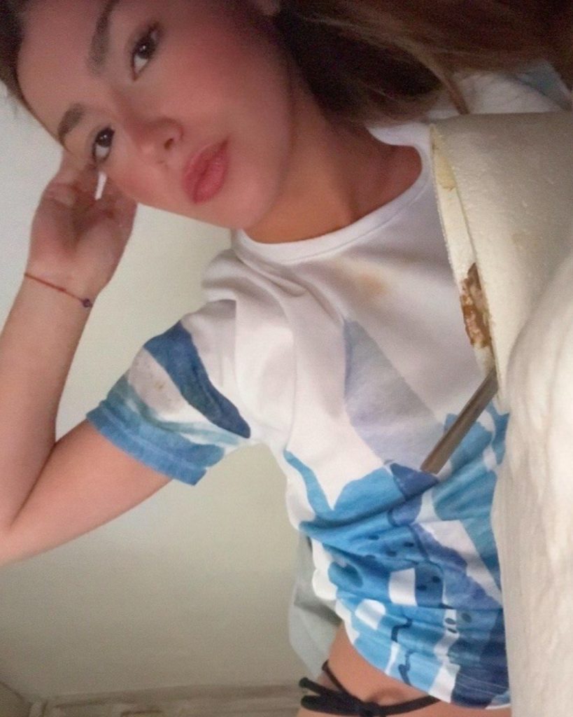 Selfie of Ava Rosabella looking toward the camera, wearing a white t-shirt with a blue abstract print, with her right hand resting gently against her temple.