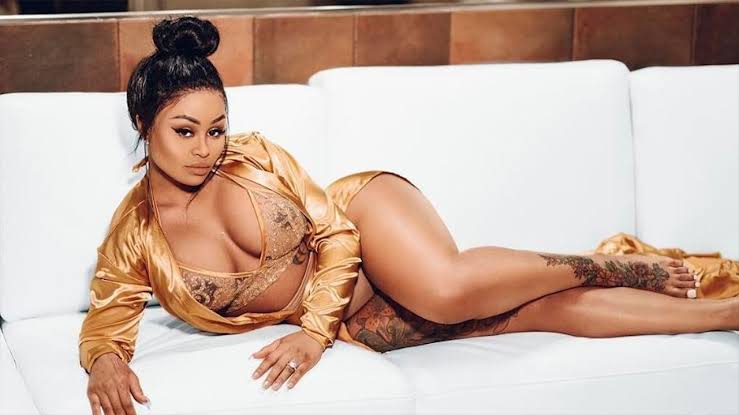 Blac Chyna, identified as a Thick OnlyFans personality, lounging on a white sofa while wearing a metallic gold bra and matching long-sleeved top.