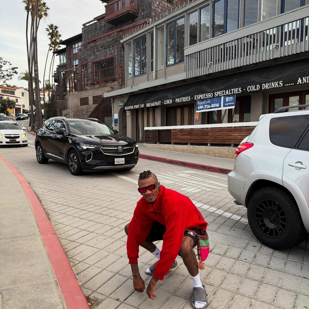 Nigerian OnlyFans creator Chimereuche-Jackson Nwokeoma crouching on a city street sidewalk wearing a red pullover jacket and sunglasses.
