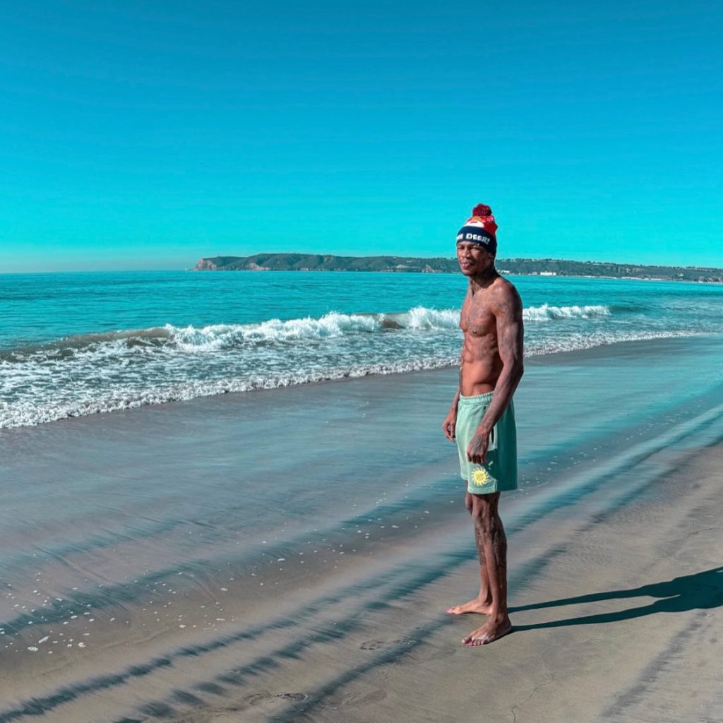 Shirtless Nigerian OnlyFans creator Chimereuche-Jackson Nwokeoma standing on a sandy beach wearing green shorts and a beanie hat.
