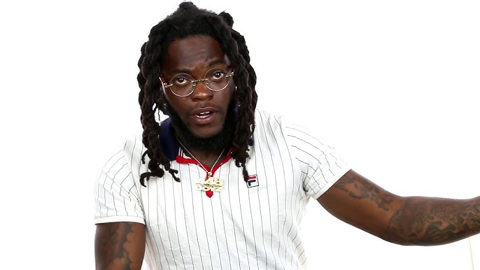 A close-up photograph of Flockatrent (@bignigerian), a leading Nigerian OnlyFans figure, wearing clear-framed glasses, a "D4L" chain, and a white pinstripe Fila polo shirt against a white background.