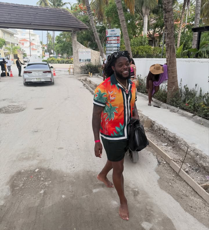 Flockatrent, a popular creator on Nigerian OnlyFans, smiling as he walks barefoot down a sandy road, wearing a bright orange tropical print short-sleeve button-up shirt and shorts.