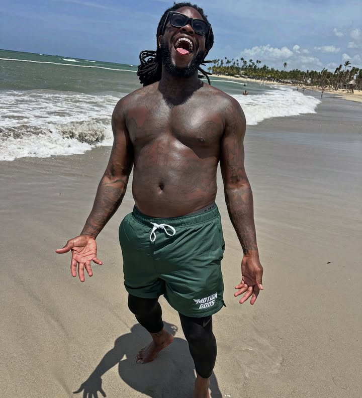 Nigerian OnlyFans creator Flockatrent poses shirtless and wide-mouthed on a sandy beach, wearing green swim trunks and sunglasses.