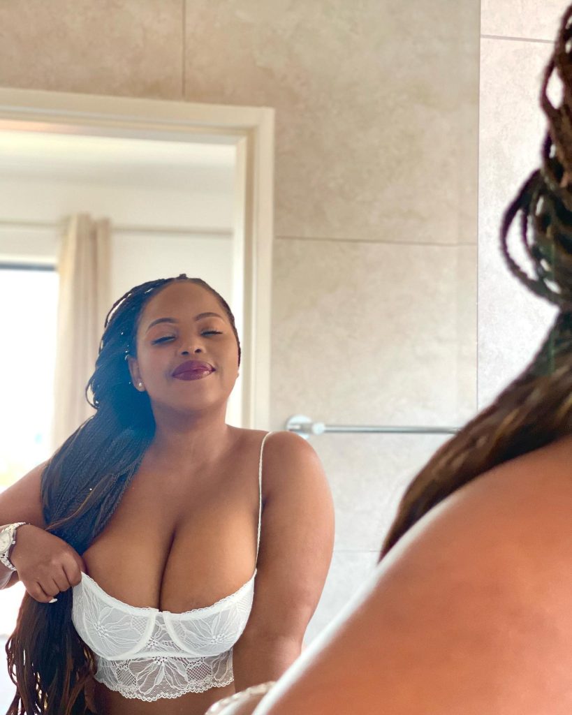 A bathroom mirror selfie of Isenako Sihlahla, a South African creator with long braids, wearing a white lace bustier and a silver watch, looking at her reflection with closed eyes.