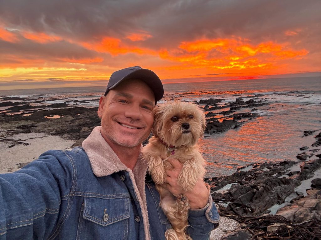 OnlyFans model Kurt Rogers holding a small dog by the ocean during a dramatic orange sunset.