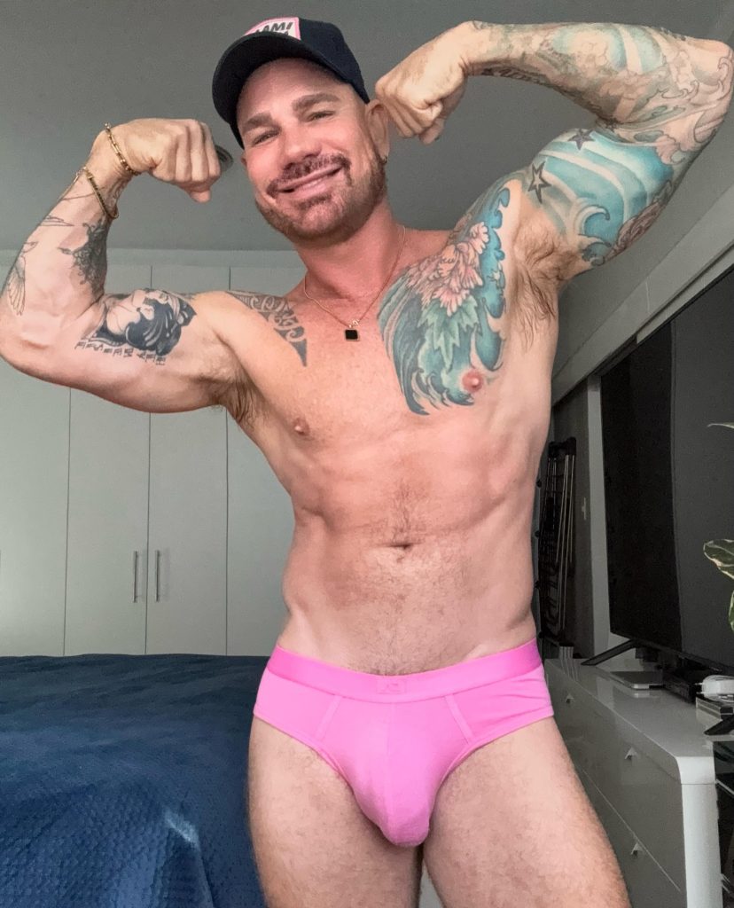 Kurt Rogers flexing his tattooed biceps in an indoor setting, wearing a black cap and pink briefs.