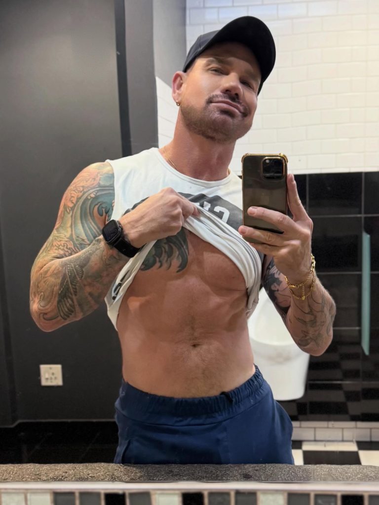 Kurt Rogers taking a mirror selfie in a public bathroom, lifting his shirt to show his abs and tattoos.