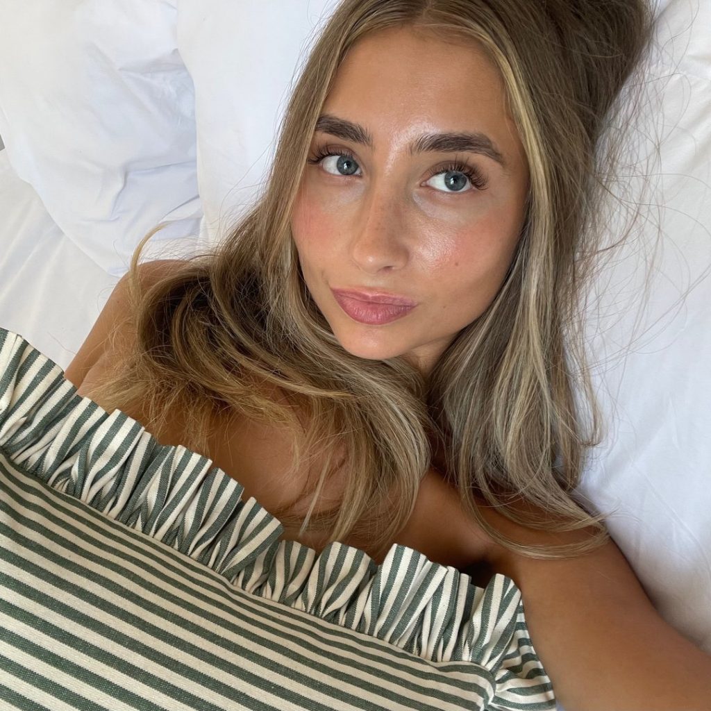 A close-up overhead selfie of Lily Phillips lying in bed against white pillows, wrapped in a green and white striped blanket, showcasing a natural and intimate look from the best GFE OnlyFans model.