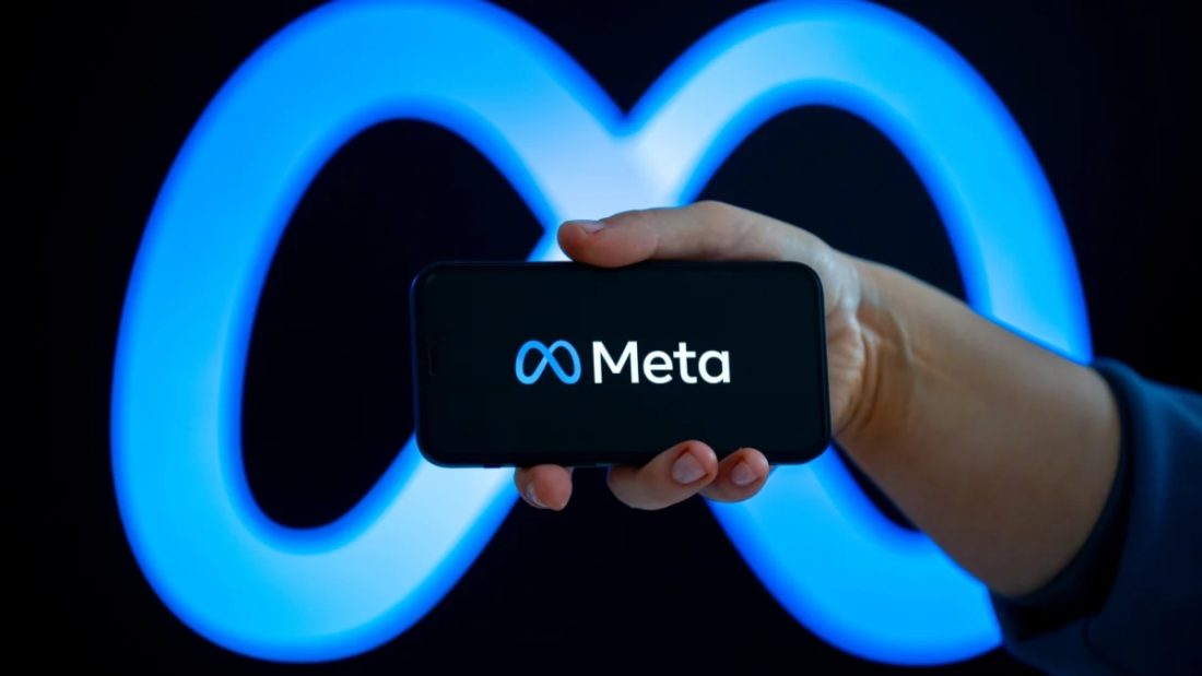 A hand holding a smartphone displaying the Meta logo on its screen against a large, glowing blue neon Meta infinity symbol background. This conceptual image represents discussions surrounding Meta AI adult content copyright policies, training data, and intellectual property issues.