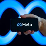 A hand holding a smartphone displaying the Meta logo on its screen against a large, glowing blue neon Meta infinity symbol background. This conceptual image represents discussions surrounding Meta AI adult content copyright policies, training data, and intellectual property issues.