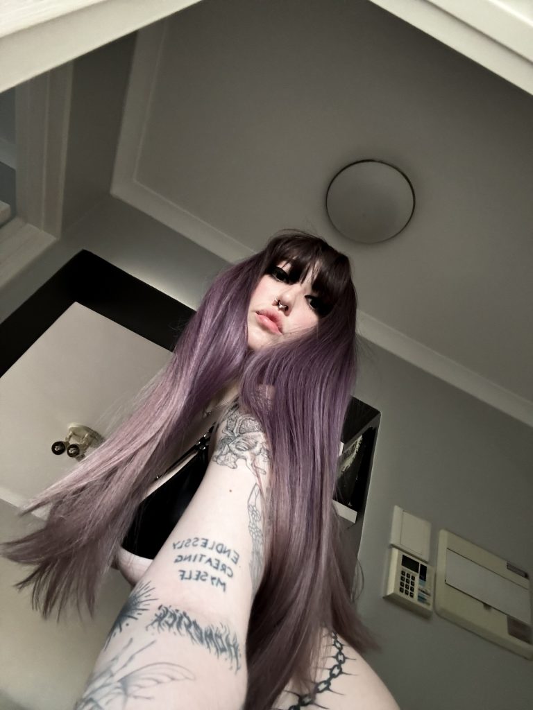 Low-angle selfie of Miya in a room with recessed lighting and a security panel, looking down with purple hair and tattoos visible.